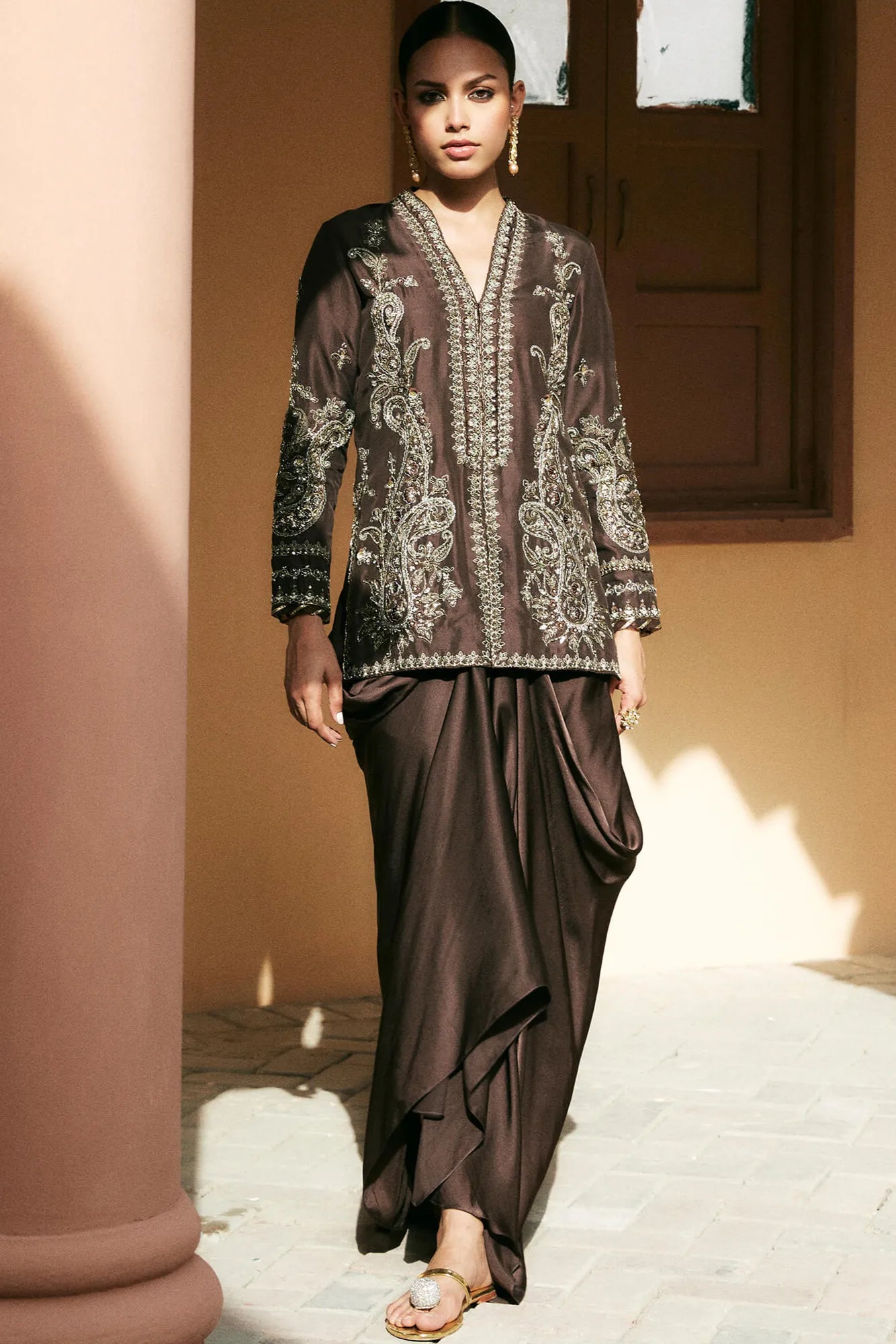 Woman wearing a brown traditional outfit with intricate patterns in an outdoor setting.