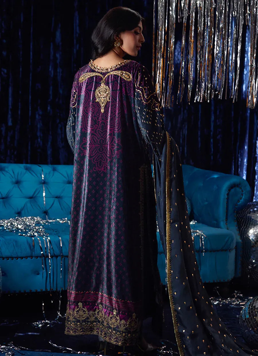 Woman in a long, dark purple embroidered dress standing in front of a blue couch with decorative drapes.