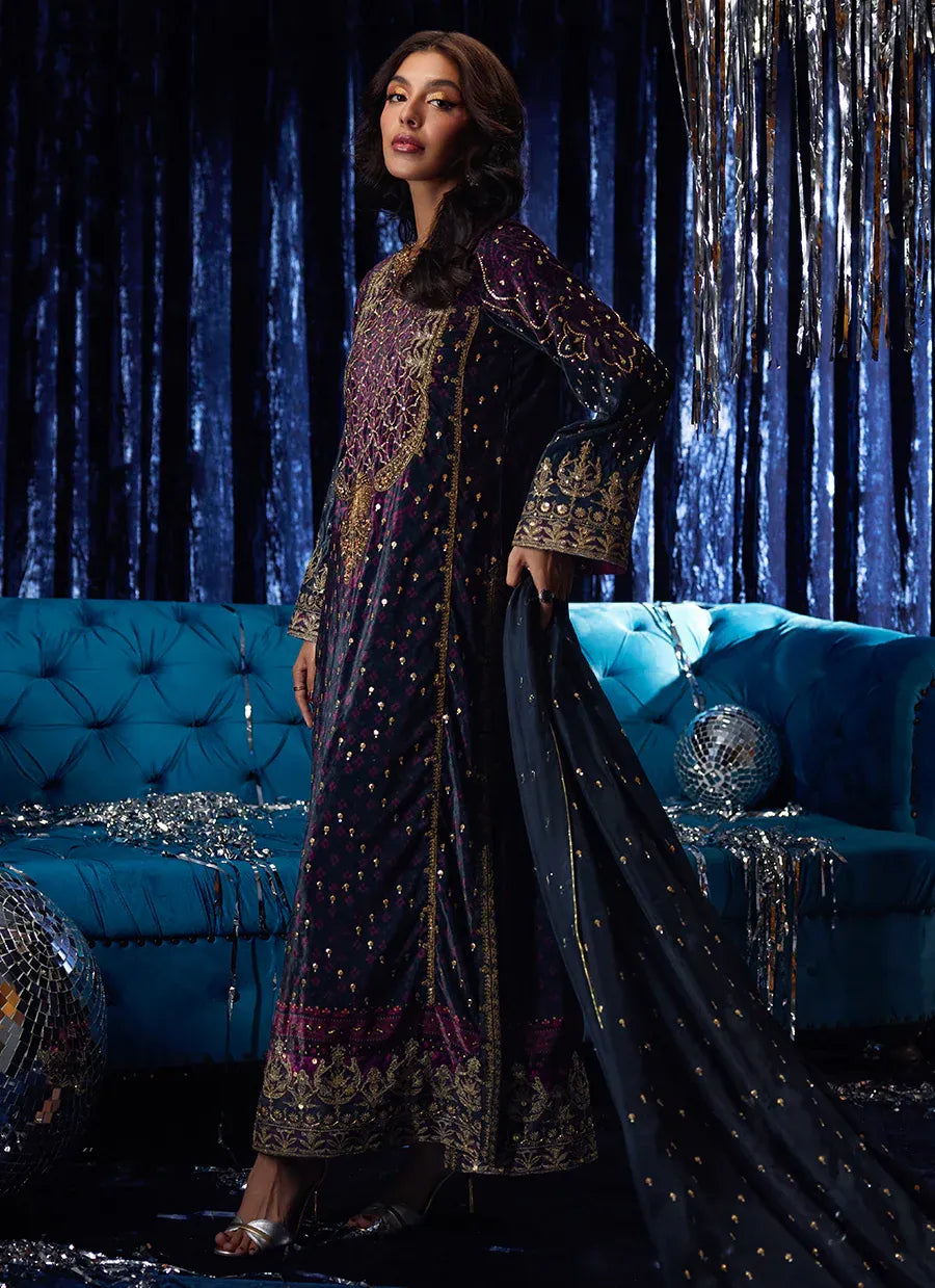 Woman in a dark blue embroidered traditional outfit standing in front of a blue couch with decorative lights.