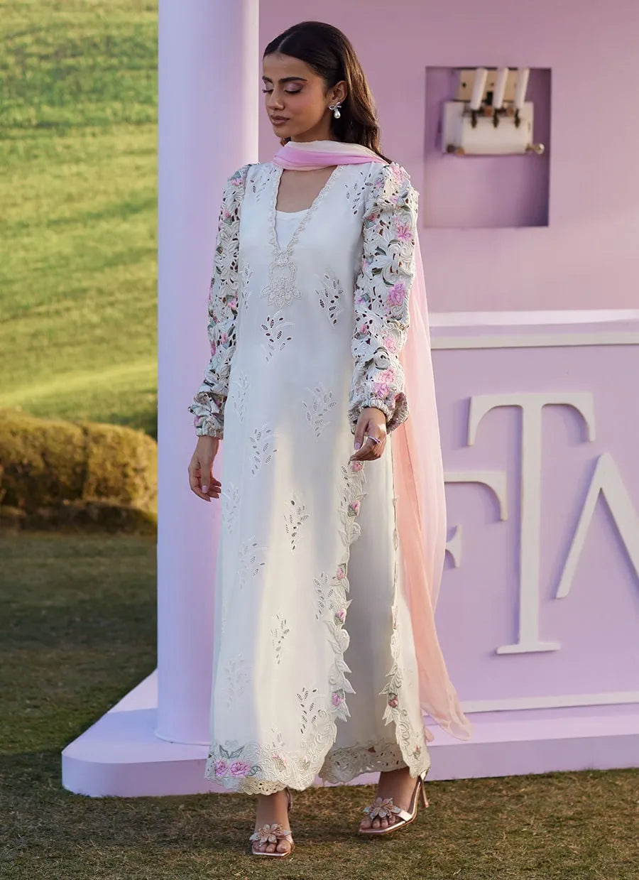 Woman in a white embroidered outfit with floral patterns standing in front of a pink and white backdrop.