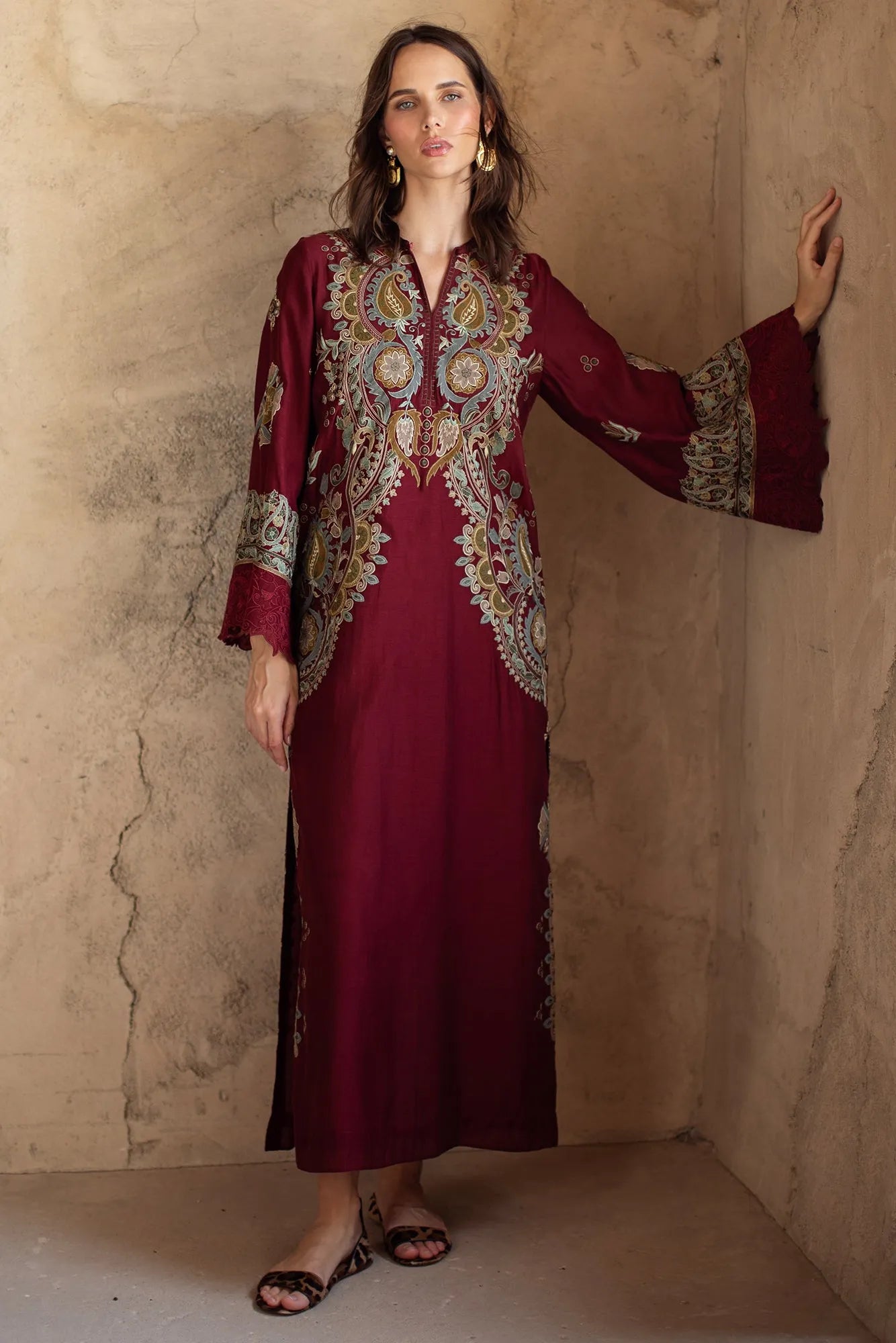 Woman wearing a burgundy dress with intricate gold embroidery against a beige wall.