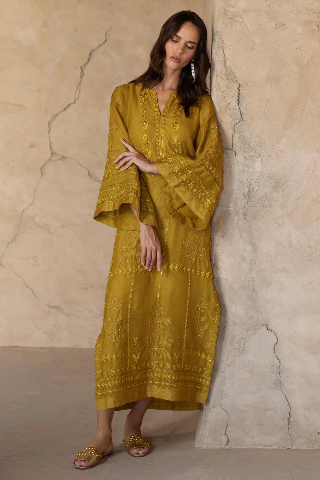 Woman wearing a mustard yellow dress with intricate patterns against a beige wall.