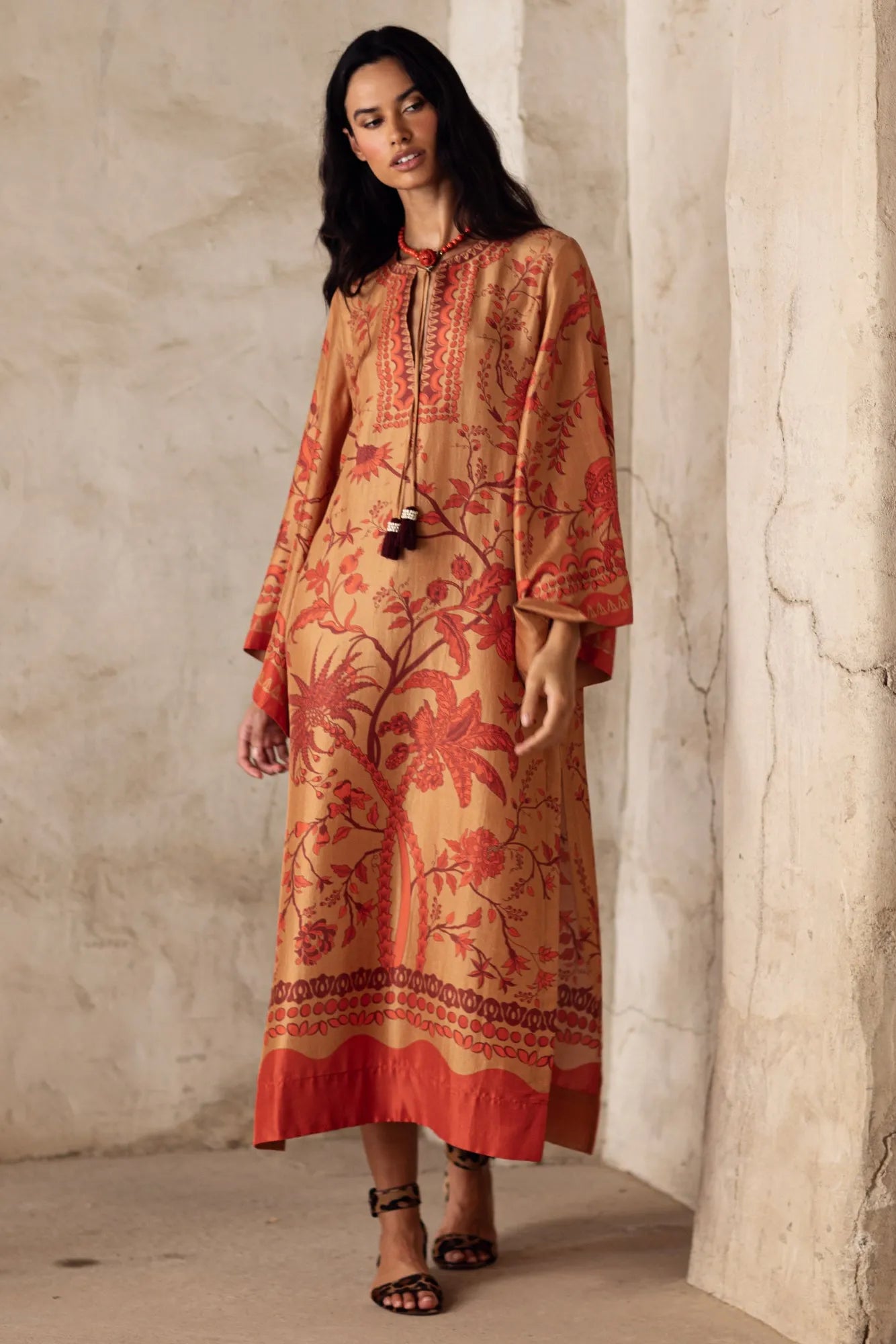 Woman wearing a long, patterned dress against a neutral wall.