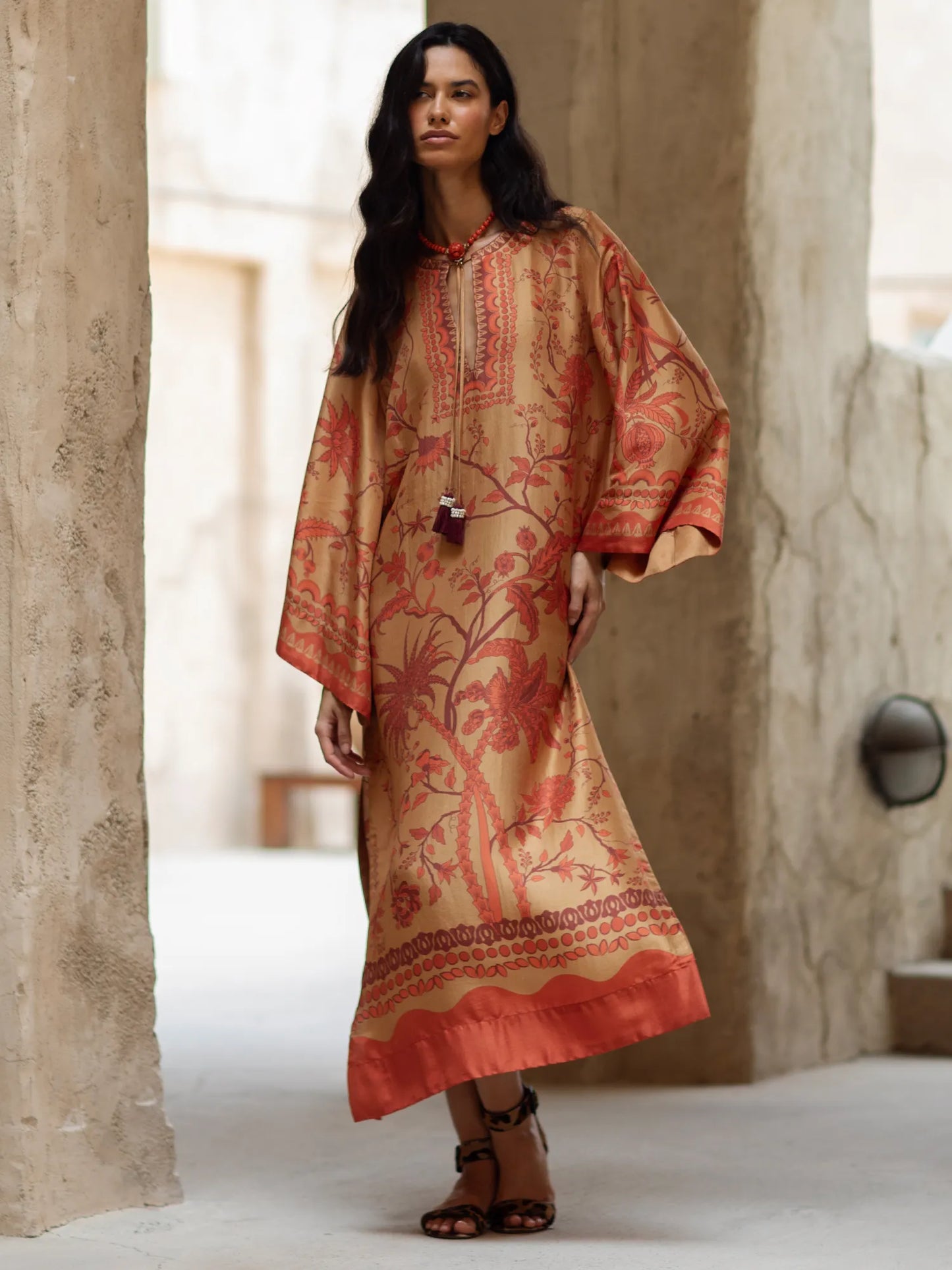 Woman wearing a patterned dress standing in a sunlit room with stone walls.