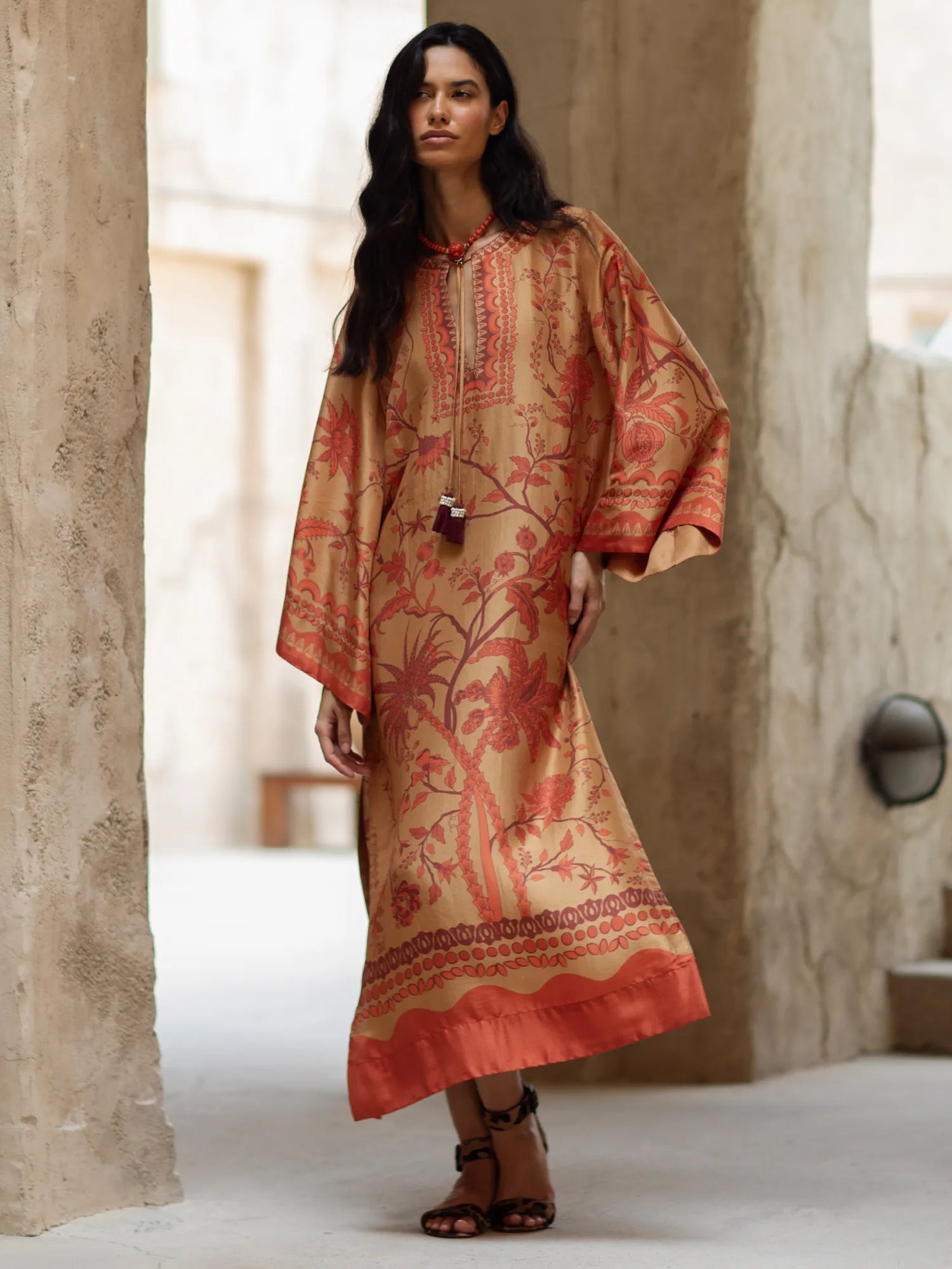 Woman wearing a patterned dress standing in a sunlit room with stone walls.