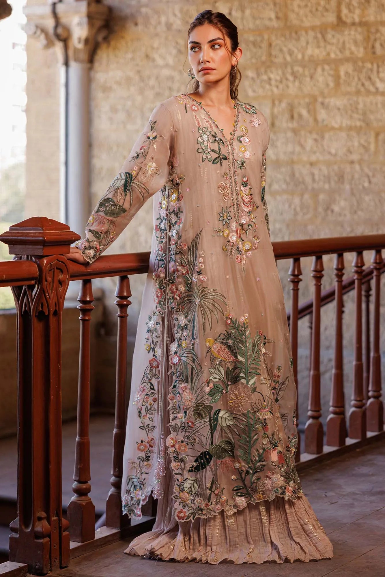 Woman in a floral embroidered dress standing on a balcony with stone walls.