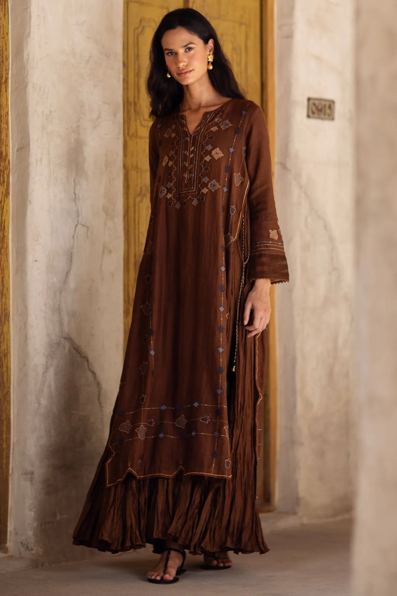 Woman wearing a long brown embroidered dress standing in a room with stone walls.