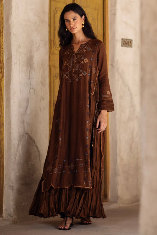 Woman wearing a long brown embroidered dress standing in a room with stone walls.