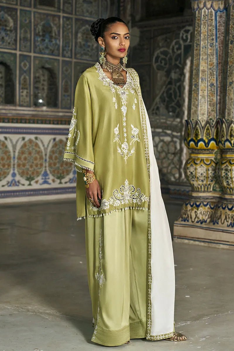 Woman in a green traditional outfit with white embroidery standing in an ornate indoor setting.