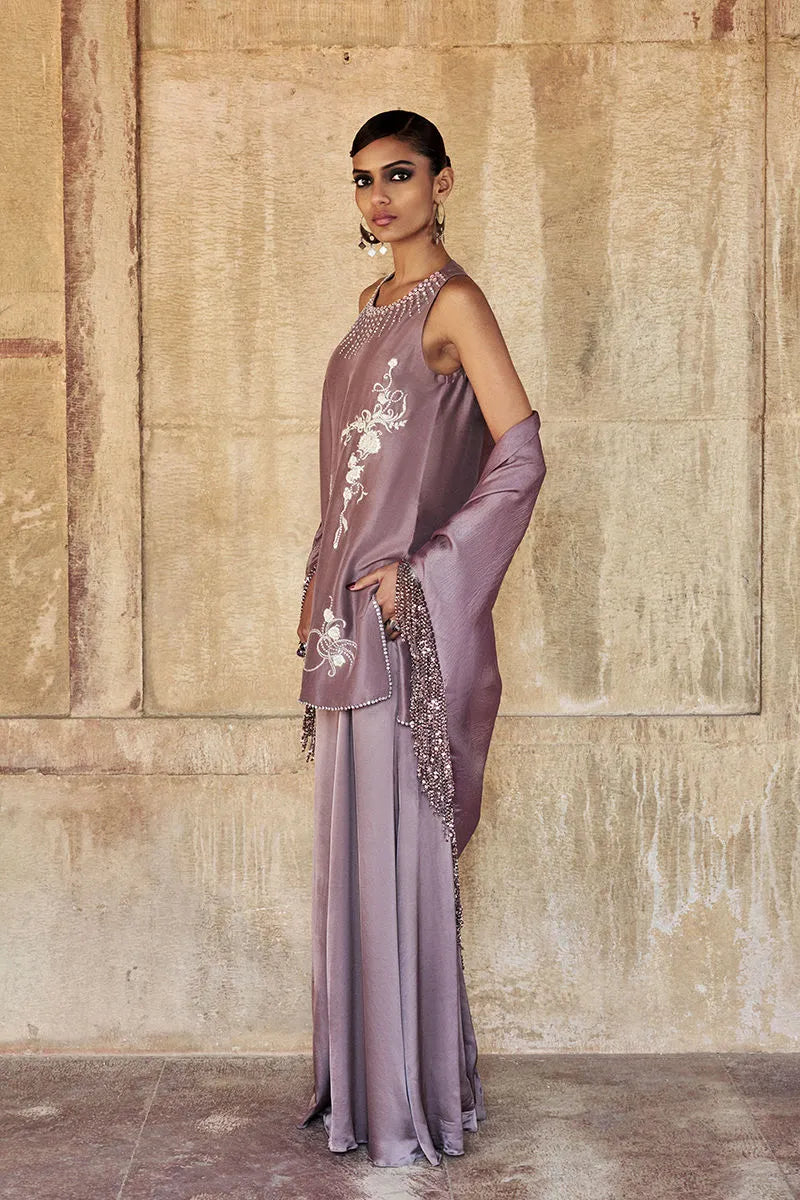 Woman in a lavender outfit with intricate detailing standing against a textured wall.