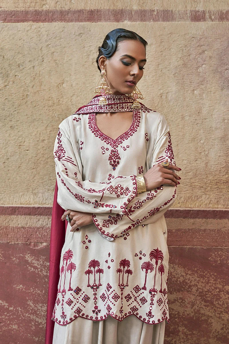 Woman wearing a cream and red embroidered traditional outfit against a textured wall.