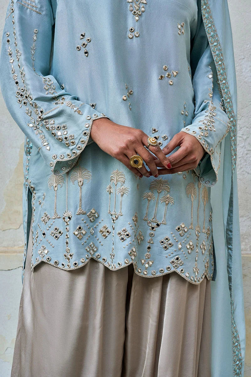 Light blue embroidered traditional outfit with beige pants against a neutral background