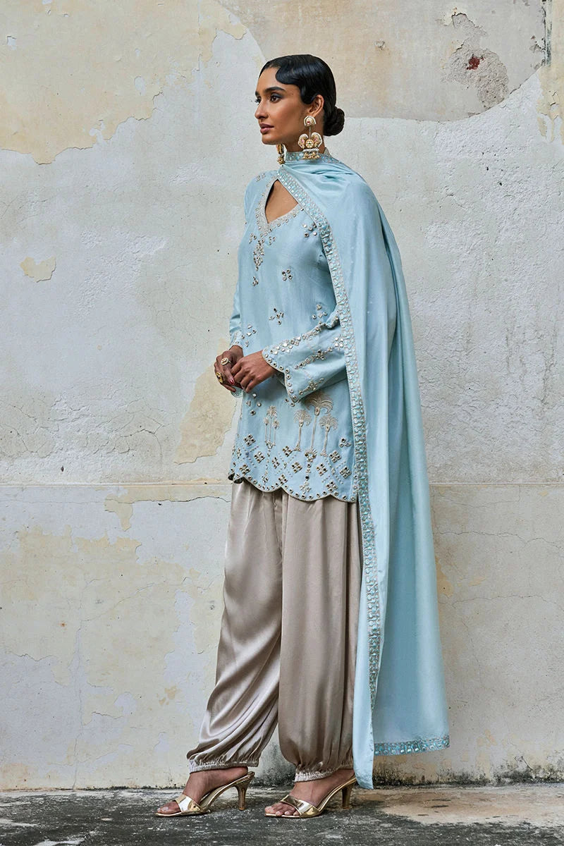Woman in traditional outfit with light blue embroidered top and beige pants against a textured wall.
