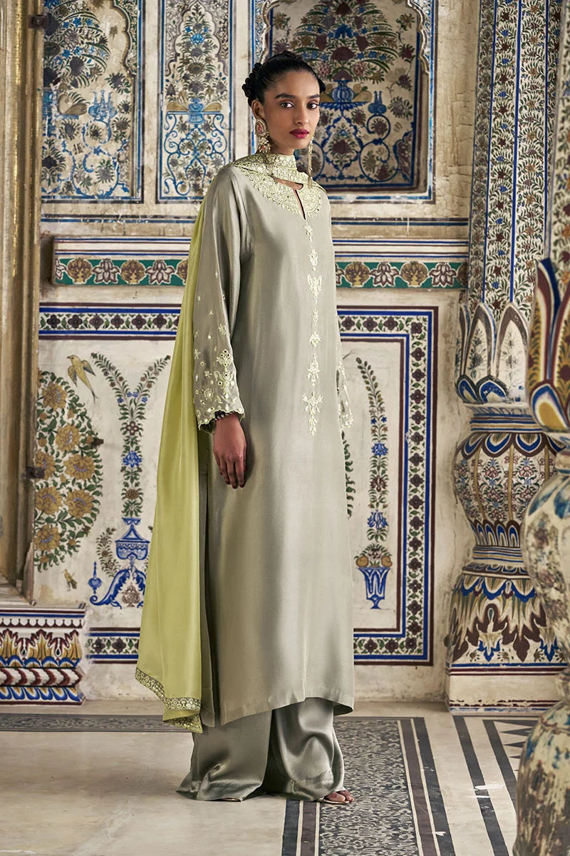Woman in a long, light-colored dress with a green shawl standing in a room with decorative tiles.