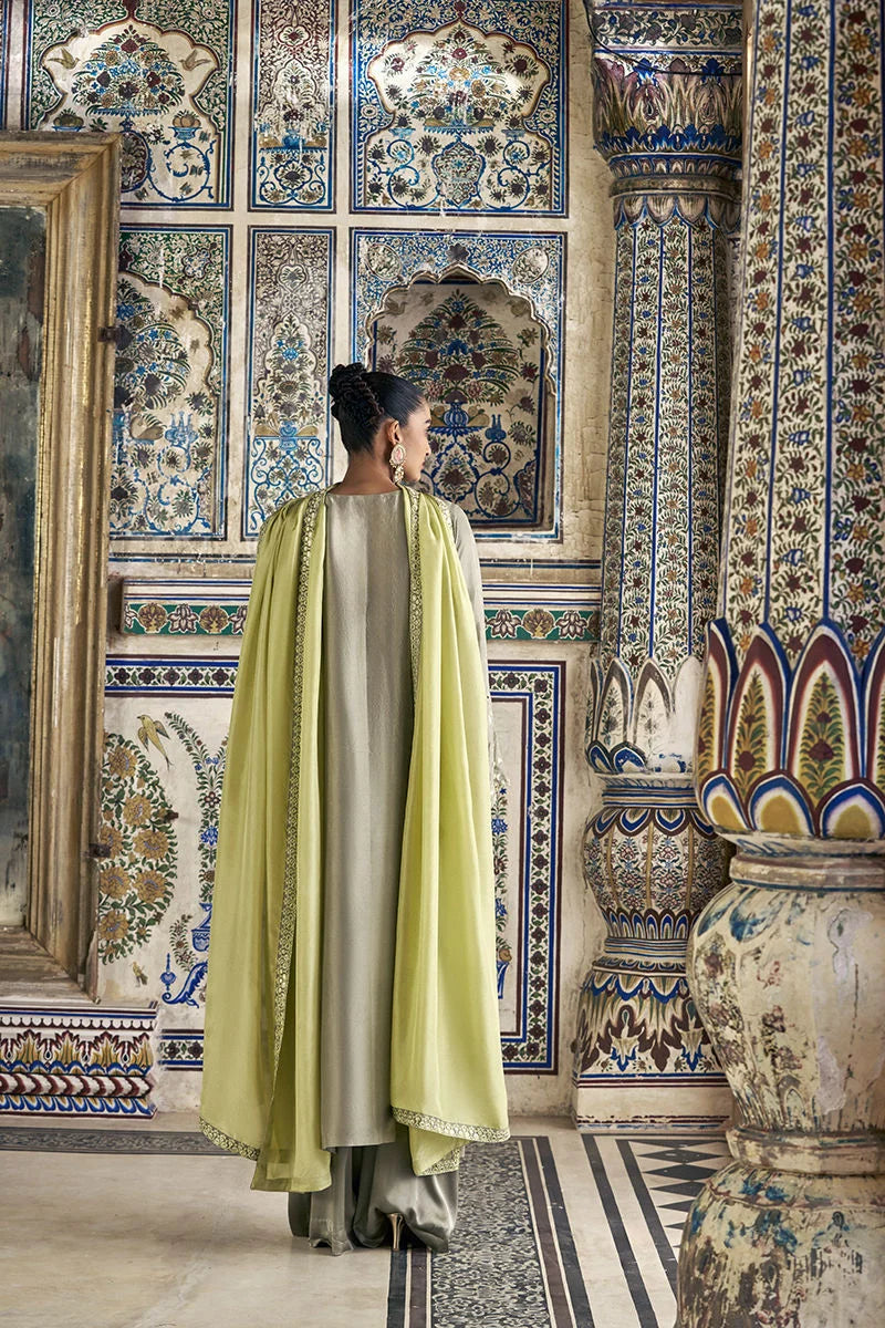 Woman in a green and white traditional outfit standing in a room with ornate wall tiles and a decorative vase.