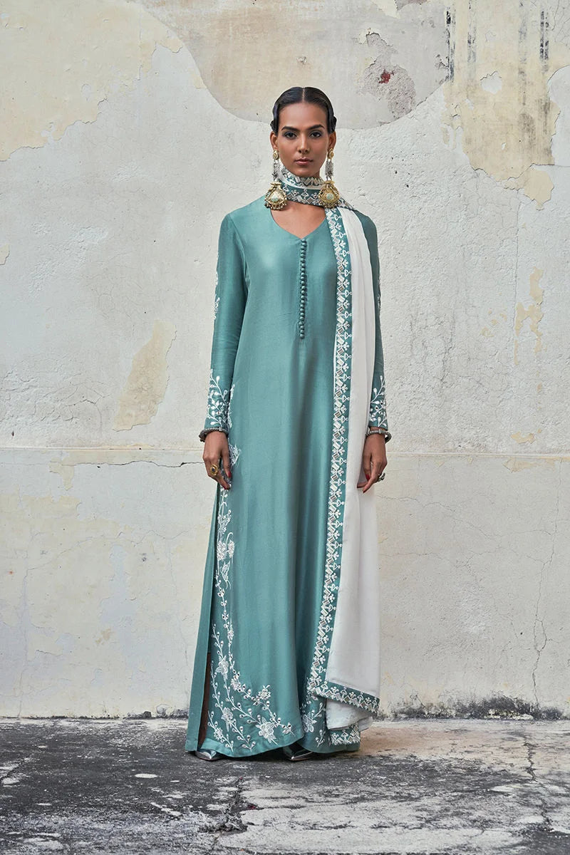 Woman wearing a teal dress with white embroidery against a textured wall.
