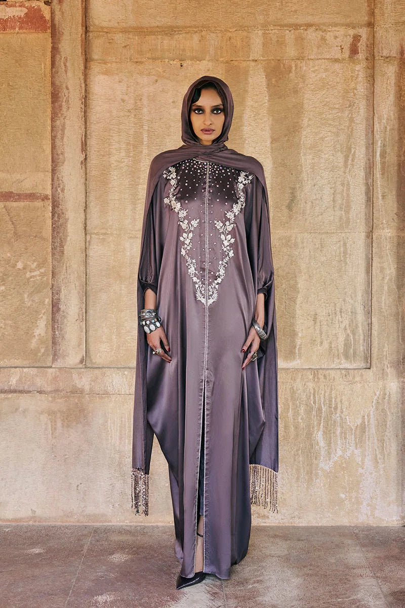 Woman wearing a long, dark purple dress with intricate white embroidery against a textured wall.