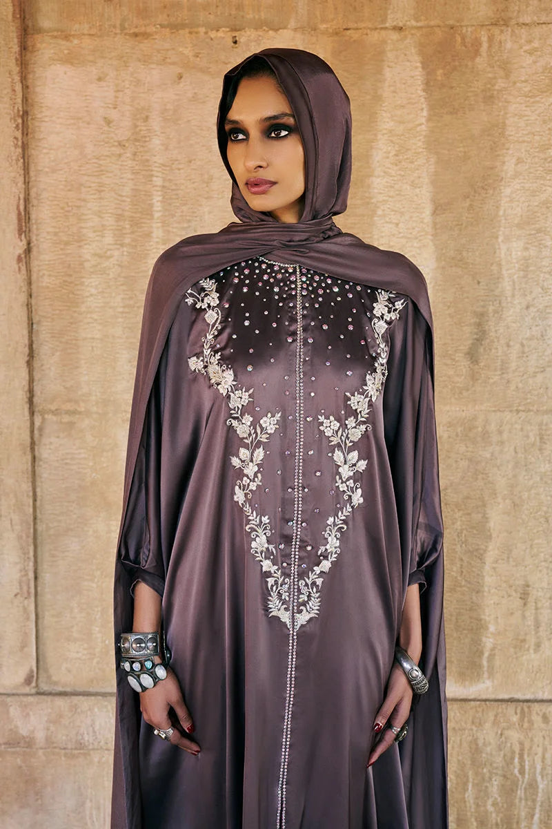 Woman wearing a dark brown abaya with white embroidery against a beige wall.