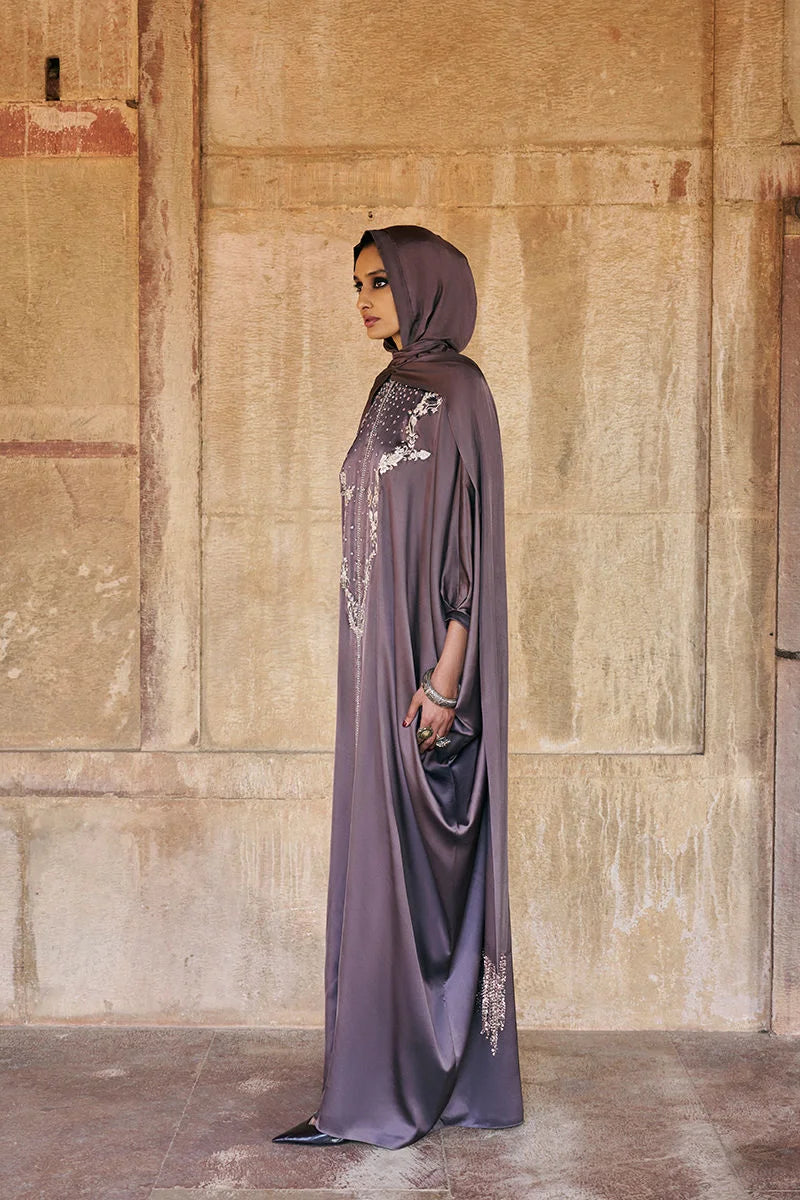 Woman in a long, light purple dress with a hood standing against a textured wall.