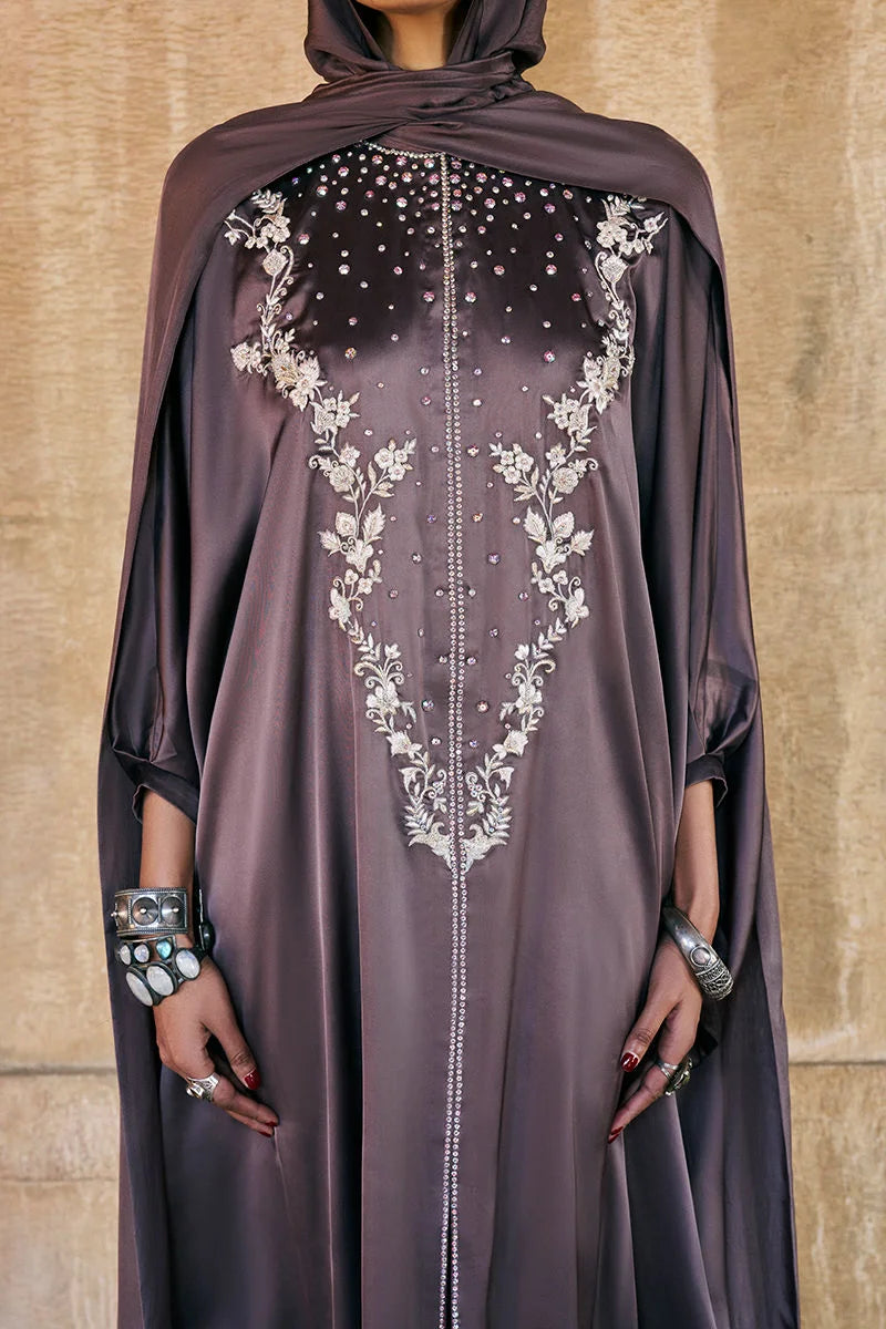 Woman wearing a dark brown abaya with intricate white embroidery against a beige wall.