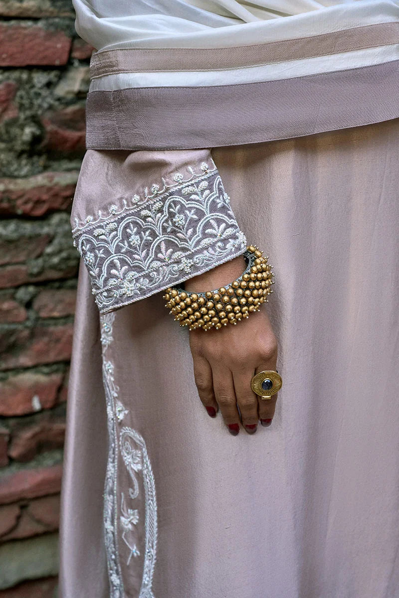 Close-up of a hand wearing gold bangles on a light pink fabric with white embroidery against a brick wall.