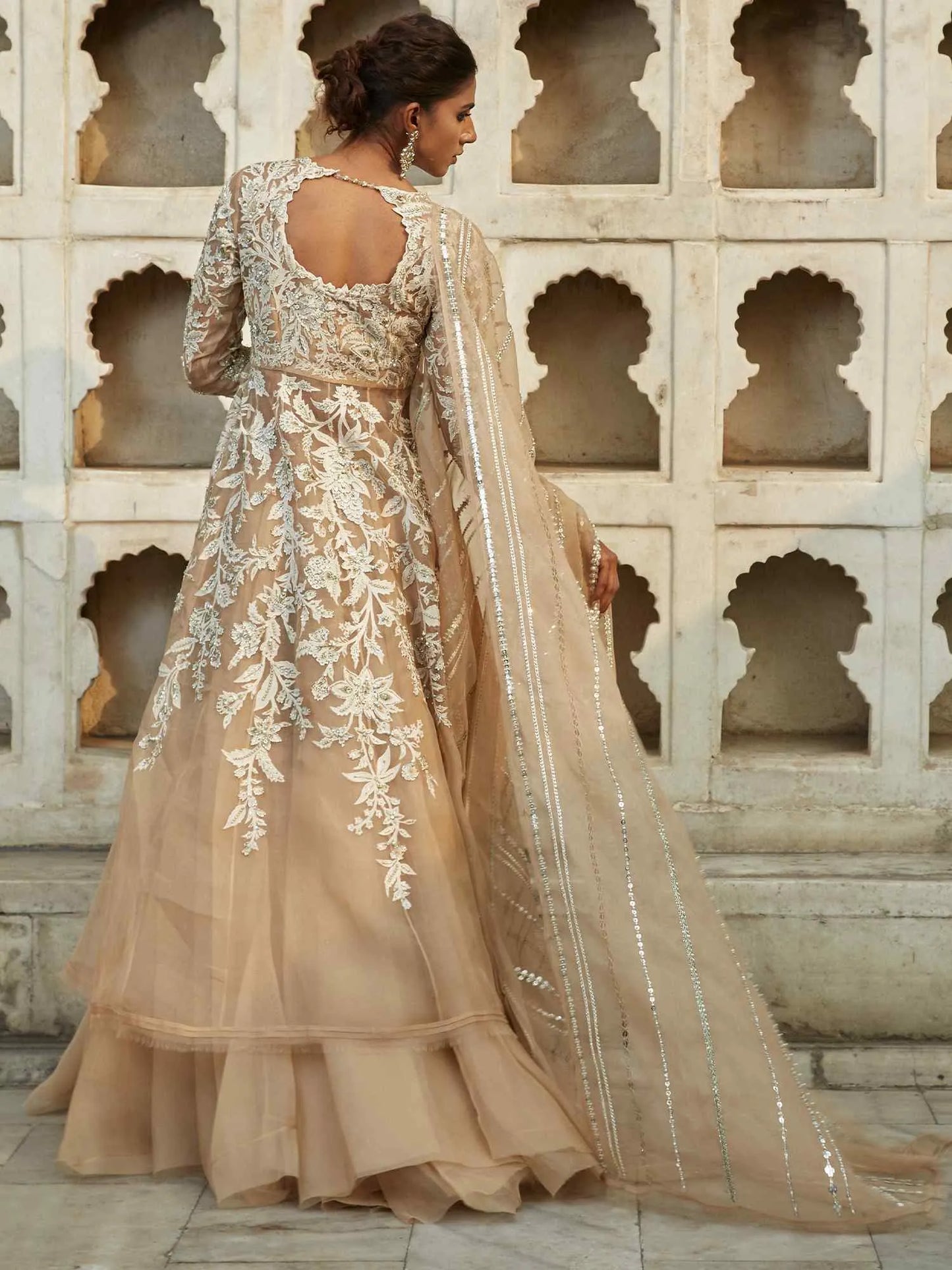 Woman in a beige embroidered traditional outfit standing in front of decorative stone walls.