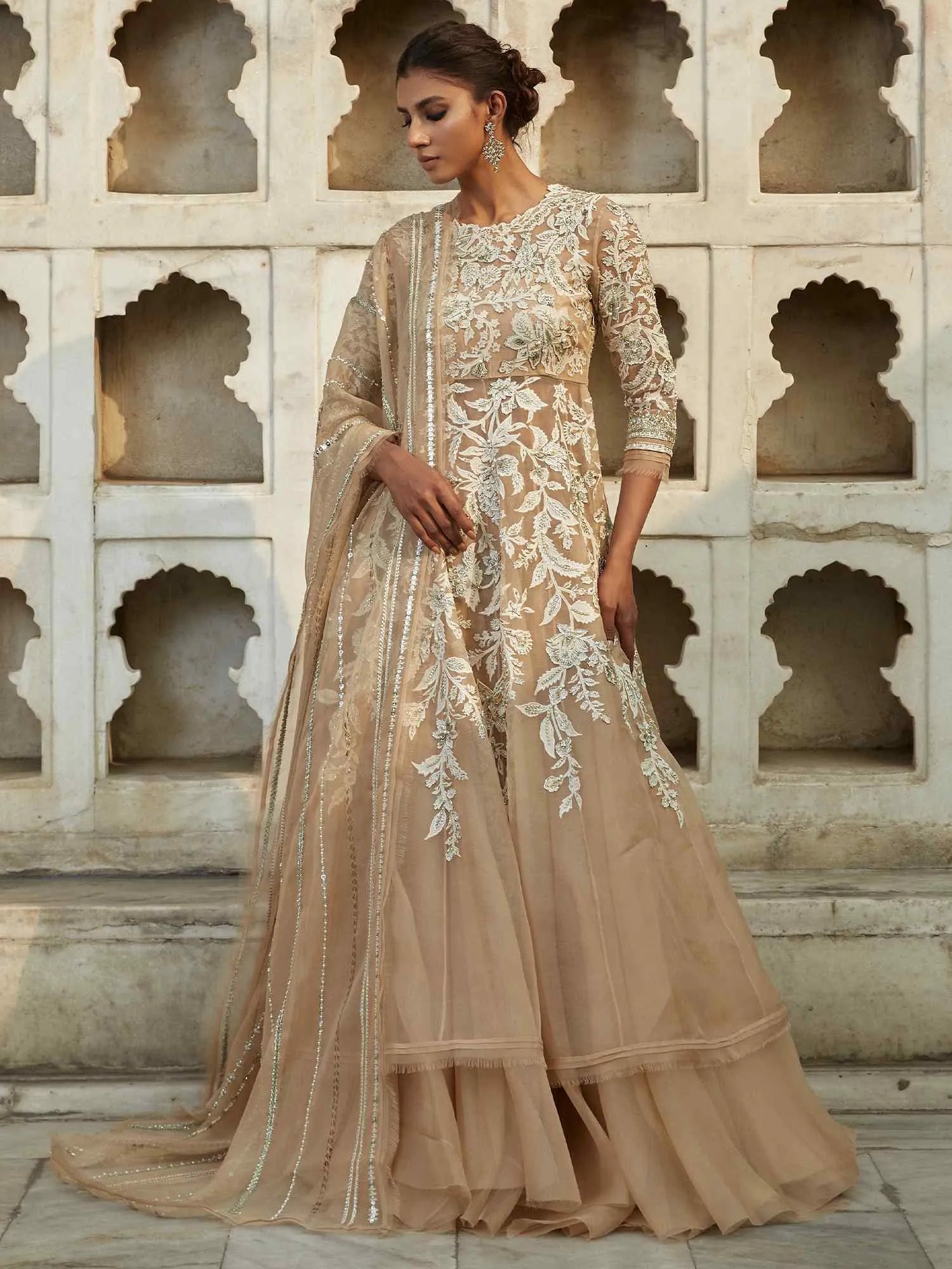 Woman wearing a beige embroidered traditional outfit in front of a stone wall with arched niches.