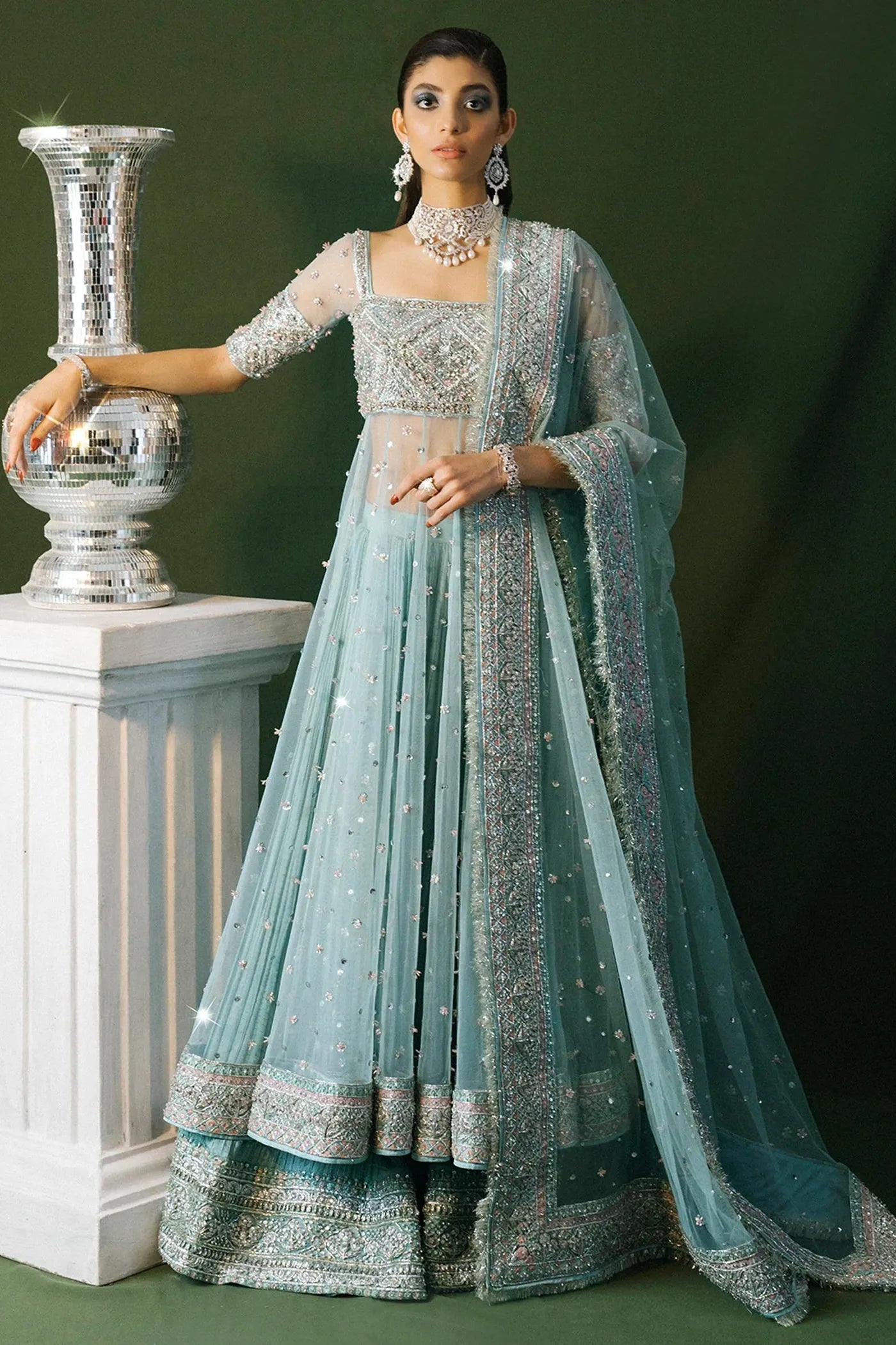Woman in a light blue and silver traditional outfit with jewelry, standing next to a decorative vase.