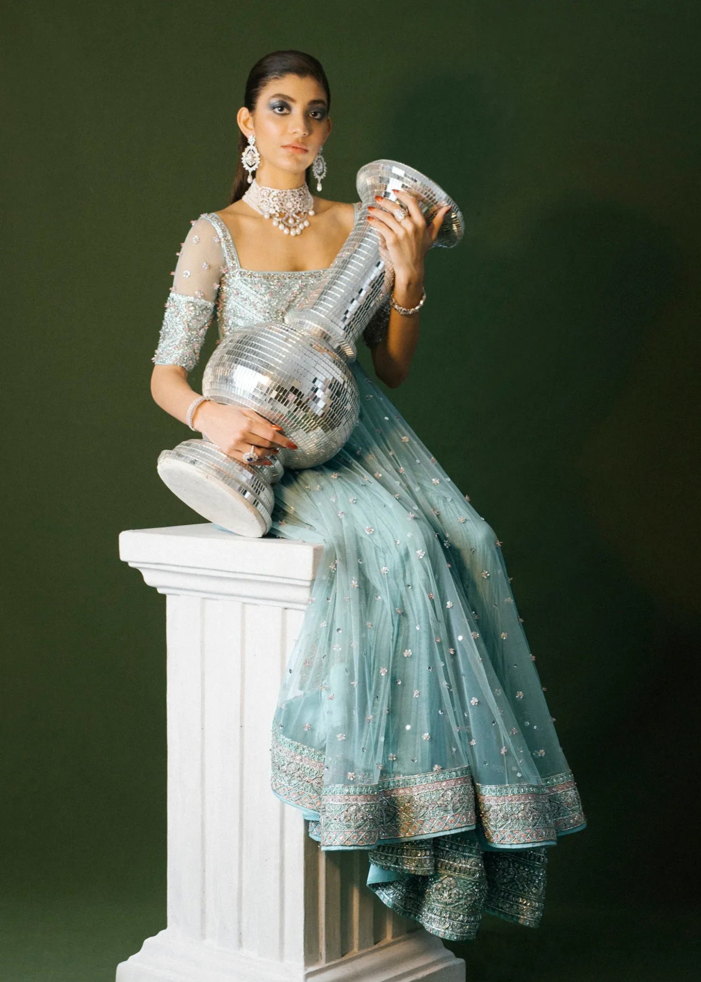 Woman in a light blue and silver traditional outfit holding a large decorative item on a white pedestal against a dark background.