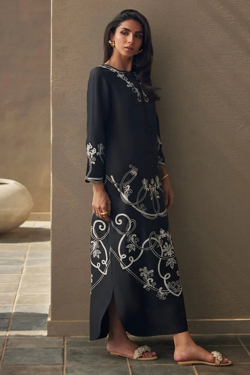 Woman wearing a black dress with white embroidery against a neutral wall.