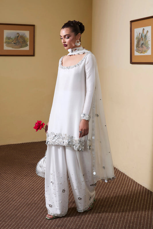 Woman in a white traditional outfit standing in a room with framed pictures on the wall.