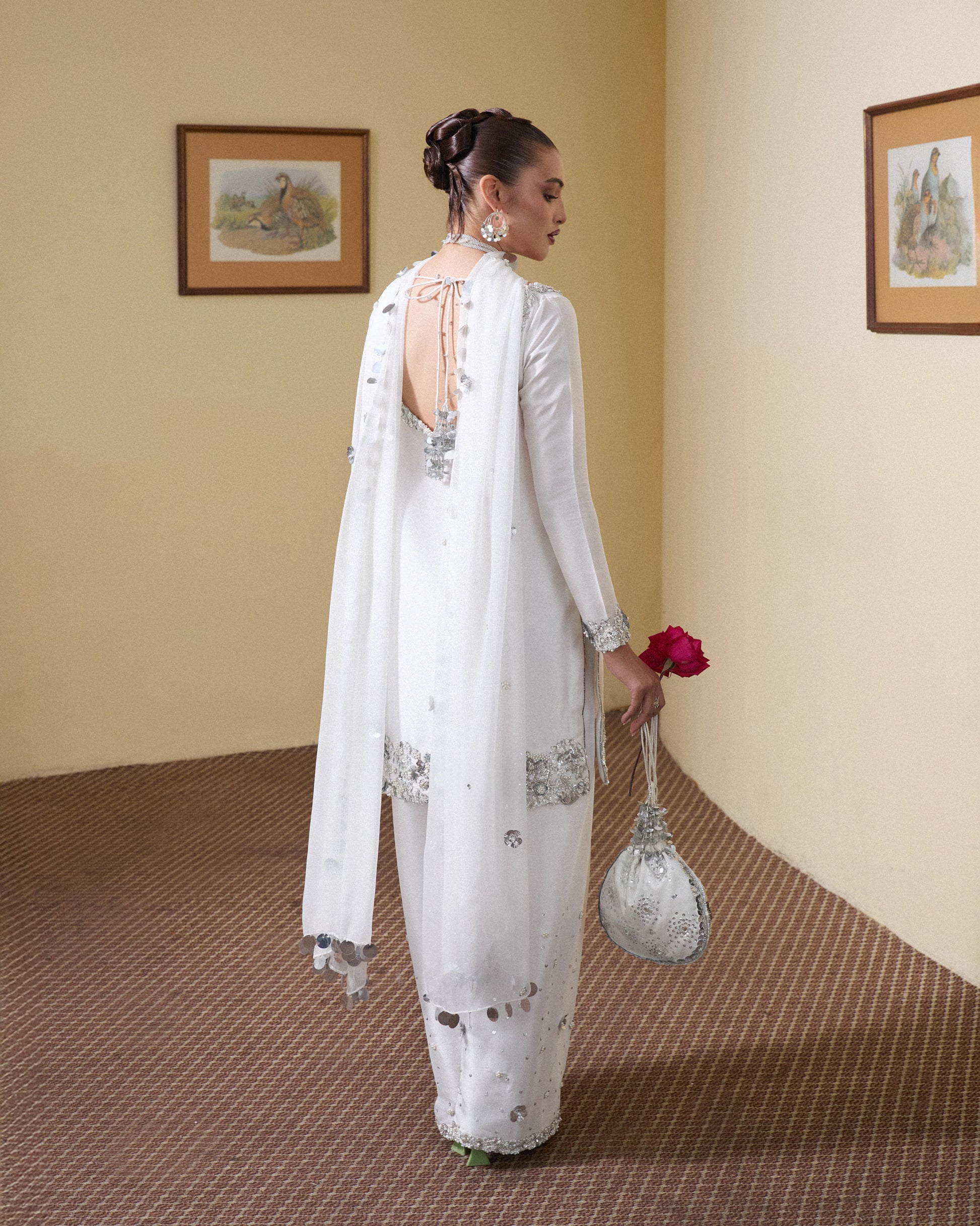 Woman in a white traditional outfit with silver embroidery, holding a red flower and a matching handbag, against a beige wall.