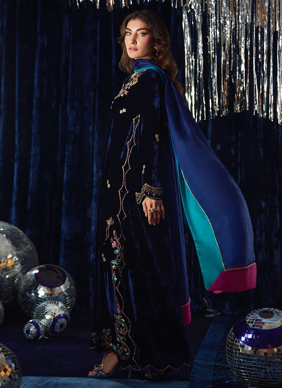 Woman in a dark blue traditional outfit with a blue and pink cape against a festive background.