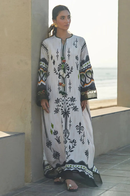 Woman wearing a white embroidered dress with black and green patterns, standing by a window with a view of the ocean.
