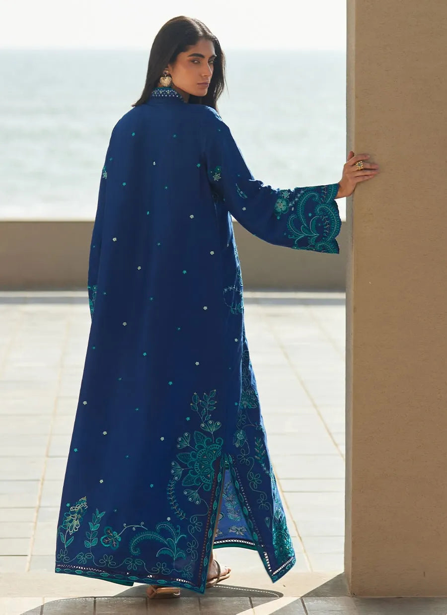 Woman wearing a blue embroidered dress standing by a window with a view of water.